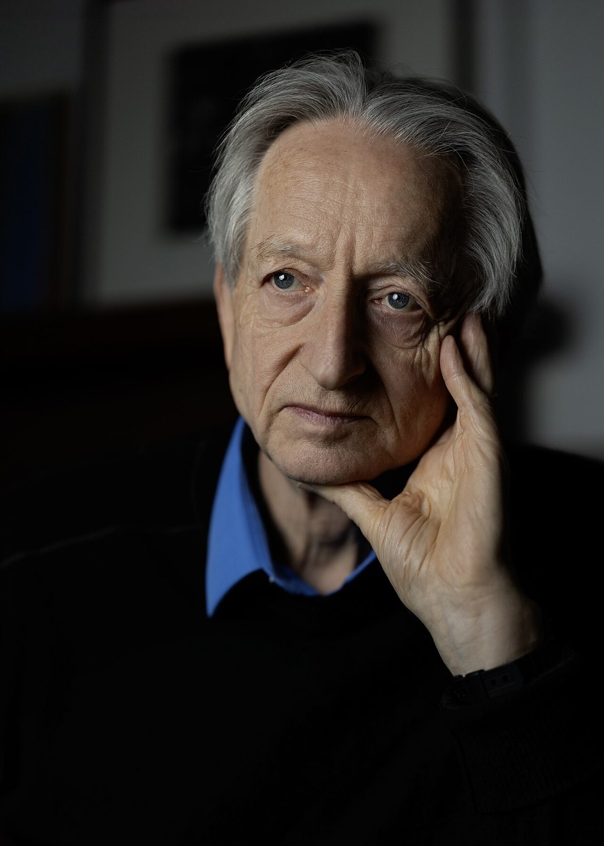 Geoffrey Hinton resting his head on one hand, in a black sweater over a blue collar, lit against a dark room