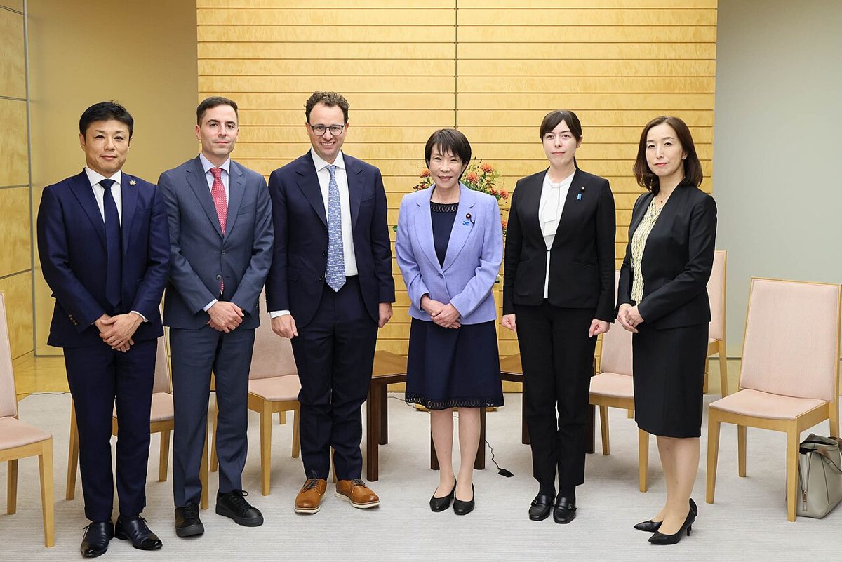 Six people in dark suits standing in a line for a photograph in a wood-paneled room, a man in glasses and a blue tie third from left