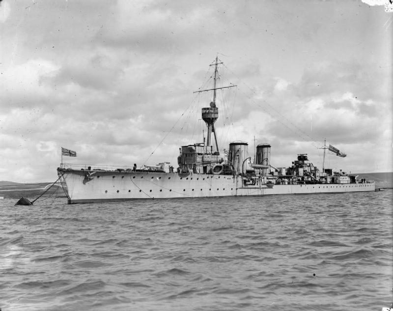 A black and white photograph of a light cruiser at anchor in choppy water, pale hull side-on, two funnels amidships and naval ensigns flying fore and aft