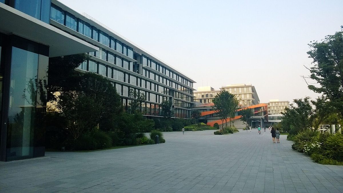 A wide paved plaza running between low glass office buildings, with young trees along both edges and a few people walking in the distance